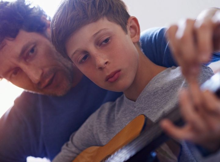 dad leans over son's shoulder to teach guitar